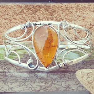 May include: A silver bangle bracelet with an amber teardrop-shaped gemstone. The bracelet has a delicate, ornate design with swirls and curves.