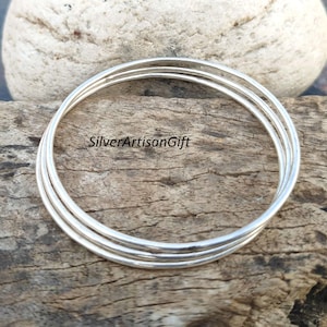 May include: Three silver bangles stacked on top of each other on a wooden surface. The bangles are smooth and shiny.