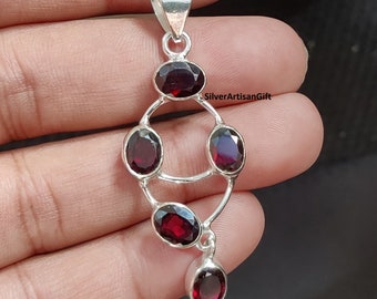 May include: A silver pendant featuring five red garnet gemstones. The pendant is shaped like a teardrop with a small round gemstone at the bottom.