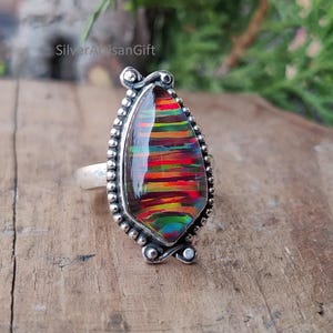 Puede incluir: Un anillo de plata con una gran gema en forma de l&aacute;grima. La gema muestra una vibrante gama de colores, incluyendo rojo, naranja, amarillo, verde y azul, en un patr&oacute;n de rayas. El anillo est&aacute; sobre una superficie de madera.