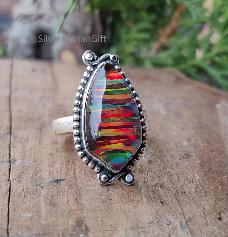 Puede incluir: Un anillo de plata con una gran gema en forma de l&aacute;grima. La gema muestra una vibrante gama de colores, incluyendo rojo, naranja, amarillo, verde y azul, en un patr&oacute;n de rayas. El anillo est&aacute; sobre una superficie de madera.