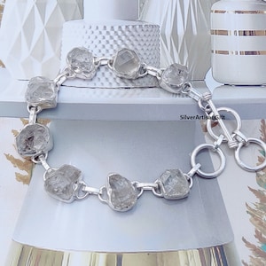 May include: A silver bracelet with a clasp closure features a chain with eight clear, faceted, rough-cut stones. The bracelet is on a white surface with a white and gold patterned background. SilverArtisanGift is printed on the surface.