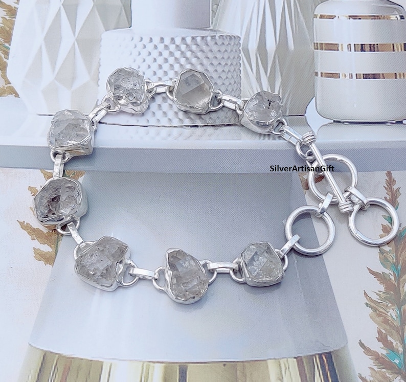 May include: A silver bracelet with a clasp closure features a chain with eight clear, faceted, rough-cut stones. The bracelet is on a white surface with a white and gold patterned background. SilverArtisanGift is printed on the surface.