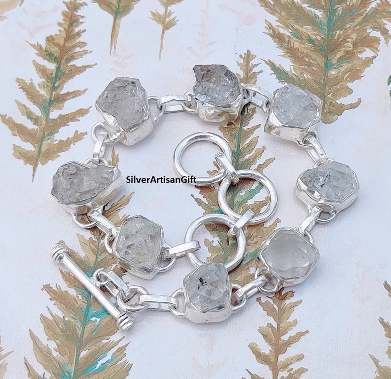 May include: A silver bracelet with a toggle clasp features faceted clear quartz stones. The bracelet is made of sterling silver and has a polished finish.  SilverArtisanGift is printed on the bracelet.