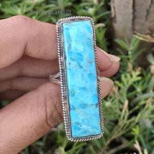 May include: A rectangular silver ring featuring a large, vibrant turquoise stone. The stone has a light blue color with darker blue veining. The ring has a twisted rope design around the stone and a simple band.