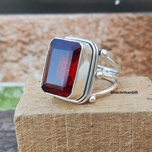 Może przedstawiać: Srebrny pierścionek z dużym, prostokątnym, czerwonym kamieniem szlachetnym. Pierścionek ma delikatną, skręconą obręcz z małymi srebrnymi koralikami.