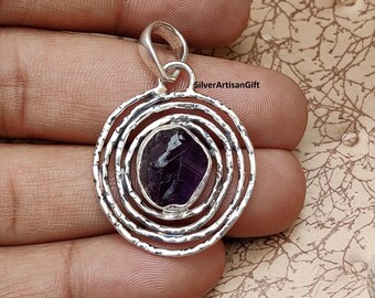 May include: A silver pendant with a circular design featuring concentric circles and a purple amethyst gemstone in the center.  The pendant has a bail for attaching to a chain.