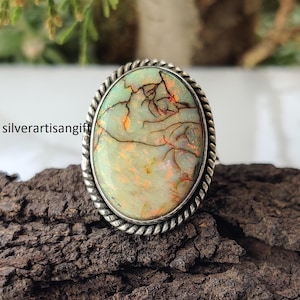 May include: Oval-shaped ring with a light green and orange gemstone, set in a silver band with a twisted rope design. The gemstone has a unique pattern of brown lines. The text "silverartisangift" is visible on the left side.