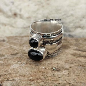 Peut inclure: Une bague en argent avec trois bandes, un charme en forme de cœur et deux pierres précieuses noires. La bague est sur une surface en bois.