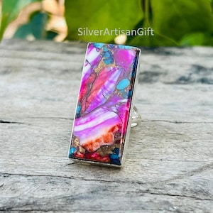 May include: A rectangular silver ring featuring a vibrant, multicolored stone. The stone displays a mix of pink, purple, orange, and blue hues with gold accents. The ring is set on a wooden surface, with the text "SilverArtisanGift" visible.