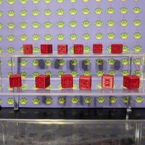 May include: A collection of red dice displayed on a clear acrylic shelf. Each die features carved markings, with the numbers and symbols visible. The background is a repeating pattern of green alien-like figures on a purple background. The dice are a vibrant red color.