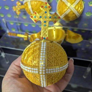 May include: A golden decorative egg with a cross on top, held in a hand. The egg is spherical with intricate patterns and white beaded accents. The cross is adorned with colorful jewels. Other similar eggs are visible in the background.