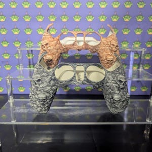 May include: A custom video game controller with a skull design. The top half is a textured brown, while the bottom half is gray with skull details. The controller sits on a clear display stand against a purple background.