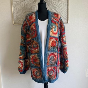 May include: A colorful, open-front crochet cardigan displayed on a black mannequin. The sweater features a patchwork design with squares in shades of blue, orange, red, and cream. The edges and trim are a solid teal color. The cardigan is a medium weight.