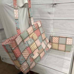 May include: A patchwork tote bag and matching wallet. The bag features a rectangular shape with long straps. The patchwork design incorporates various fabrics in shades of pink, blue, and beige. The wallet mirrors the bag's design.