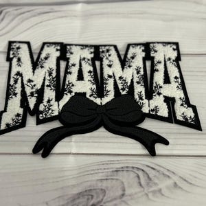 May include: Black and white fabric patch with the word "MAMA" in a floral pattern with a black bow in the centre.
