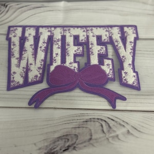 Peut inclure: Applique en feutre violet avec le mot "WIFEY" en lettres blanches avec un motif floral. Un nœud violet est centré sous le mot.
