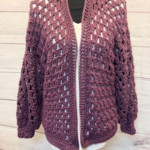 May include: A plum-colored, open-front crochet cardigan with long sleeves. The handmade sweater features a diamond-shaped openwork design and a shawl collar. The cardigan is displayed on a black mannequin, showcasing its texture and style.