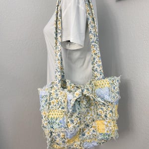 May include: A patchwork tote bag with a floral pattern in yellow, blue, and white. The bag has long straps and a ruffled texture. The bag is displayed on a mannequin. The bag is handmade.