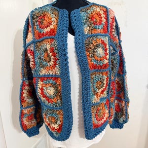 May include: A long-sleeved cardigan constructed from multi-colored crochet granny squares in shades of orange, red, blue, and cream. The squares are joined by a solid blue yarn border that also forms the cuffs, neckline, and front opening edges.