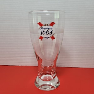 Może przedstawiać: Przezroczysty szklany kufel do piwa z rozszerzaną górą i zaokrągloną podstawą. Na szkle widnieje napis "Kronenbourg 1664" w kolorze czerwono-białym. Szklanka jest pusta i stoi na czerwonej powierzchni.