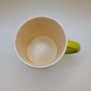 May include: A ceramic coffee mug with a speckled interior and a bright green handle. The mug's interior is a light beige color, and the exterior is white with a green accent. The mug is empty and sits on a white surface.