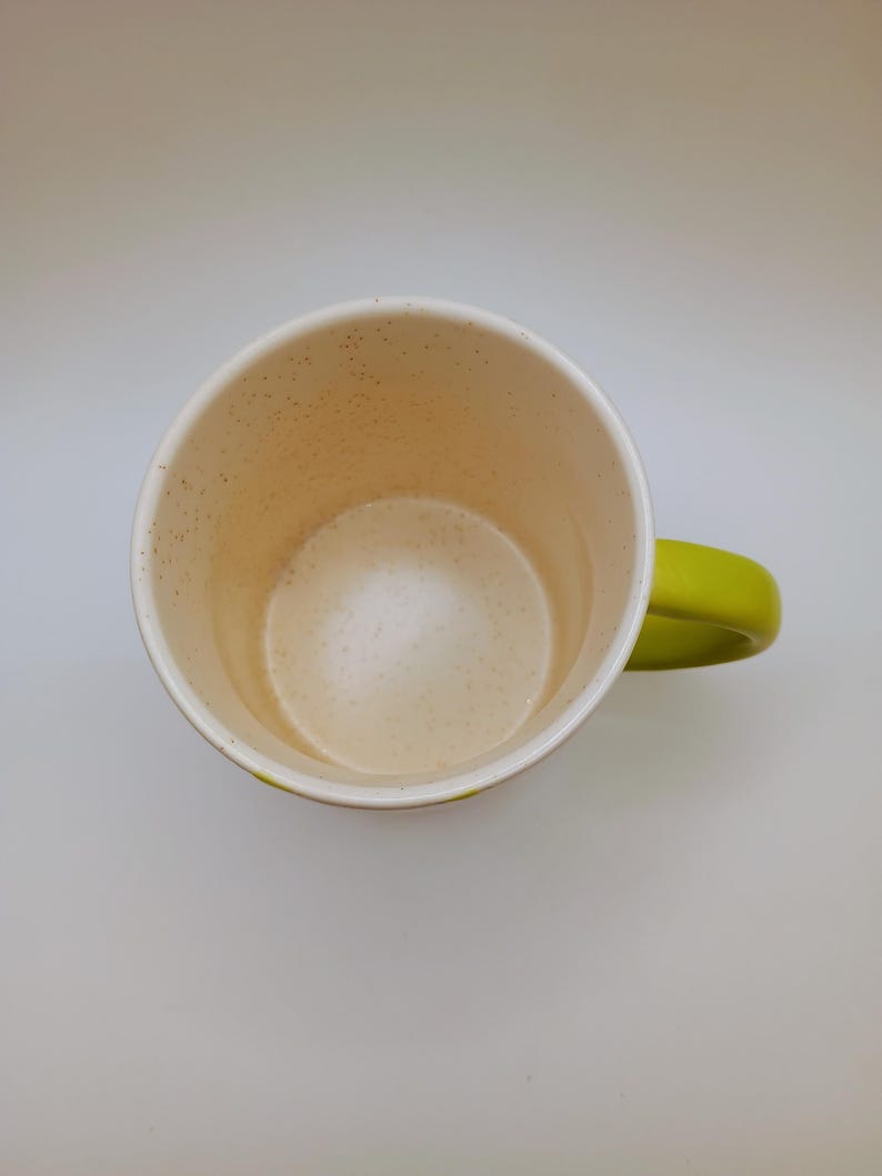 May include: A ceramic coffee mug with a speckled interior and a bright green handle. The mug's interior is a light beige color, and the exterior is white with a green accent. The mug is empty and sits on a white surface.