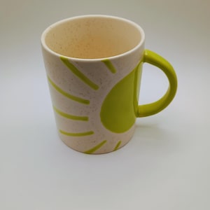 May include: A ceramic mug with a speckled off-white exterior and a bright green handle. The mug features a sunburst design in green on the side. The interior is off-white. This mug is suitable for hot or cold beverages.