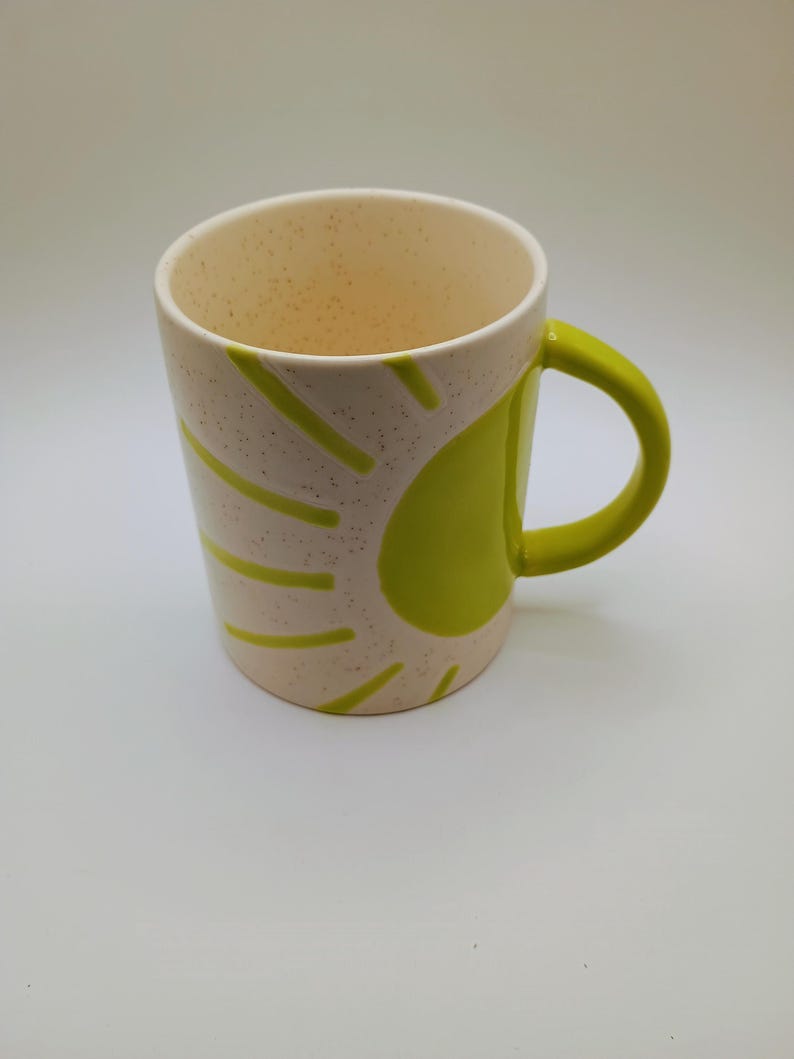 May include: A ceramic mug with a speckled off-white exterior and a bright green handle. The mug features a sunburst design in green on the side. The interior is off-white. This mug is suitable for hot or cold beverages.