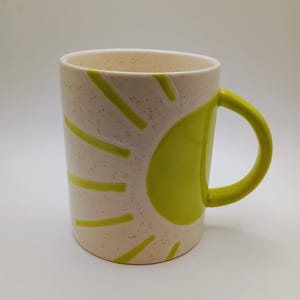 May include: A ceramic mug with a cream-colored speckled finish. The mug features a bright green sun design on the side and a matching green handle. The mug is cylindrical and suitable for hot or cold beverages.