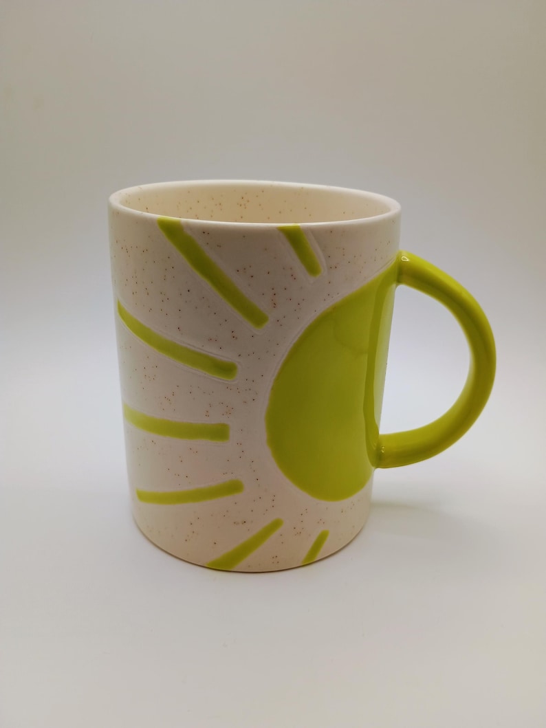 May include: A ceramic mug with a cream-colored speckled finish. The mug features a bright green sun design on the side and a matching green handle. The mug is cylindrical and suitable for hot or cold beverages.