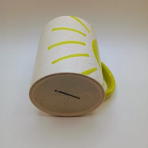 May include: A white ceramic mug with a speckled finish and a bright yellow handle. The mug features a sunburst design in yellow. The bottom of the mug has the text "by ANTHROPOLOGIE".