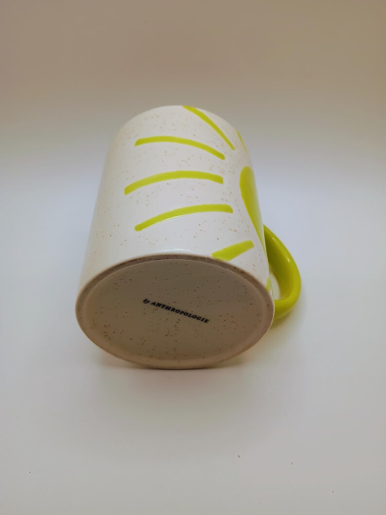 May include: A white ceramic mug with a speckled finish and a bright yellow handle. The mug features a sunburst design in yellow. The bottom of the mug has the text "by ANTHROPOLOGIE".