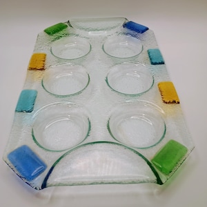 May include: Clear glass serving tray with eight circular indentations for cups or small bowls. The tray has a rectangular shape with colorful glass accents in blue, green, and yellow. The glass has a textured surface.