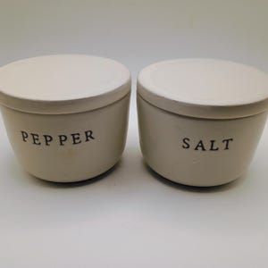 May include: Two off-white ceramic salt and pepper containers with matching lids. The word "PEPPER" is printed in black on one container, and "SALT" on the other. The containers have a simple, classic design.