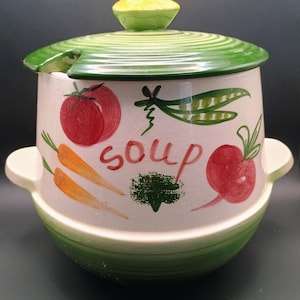 May include: A ceramic soup tureen with a lid and handles. The tureen is white with green and red painted vegetables, and the word "soup" in red. The lid has a green rim and a yellow knob.