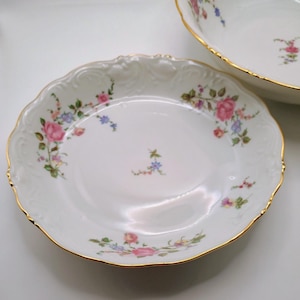 May include: Two white porcelain plates with a floral design. The plates have a scalloped edge and a gold rim. The floral design includes pink, blue, and yellow flowers with green leaves. One plate is in the foreground, and the other is partially visible in the background.