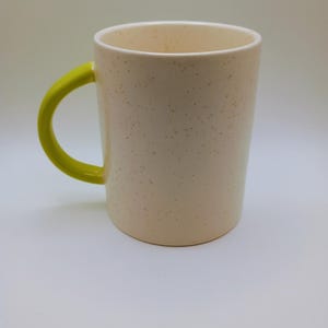 May include: A speckled cream-colored ceramic mug with a bright yellow handle. The mug has a cylindrical shape and a simple design, suitable for coffee, tea, or other beverages. The interior of the mug is white.
