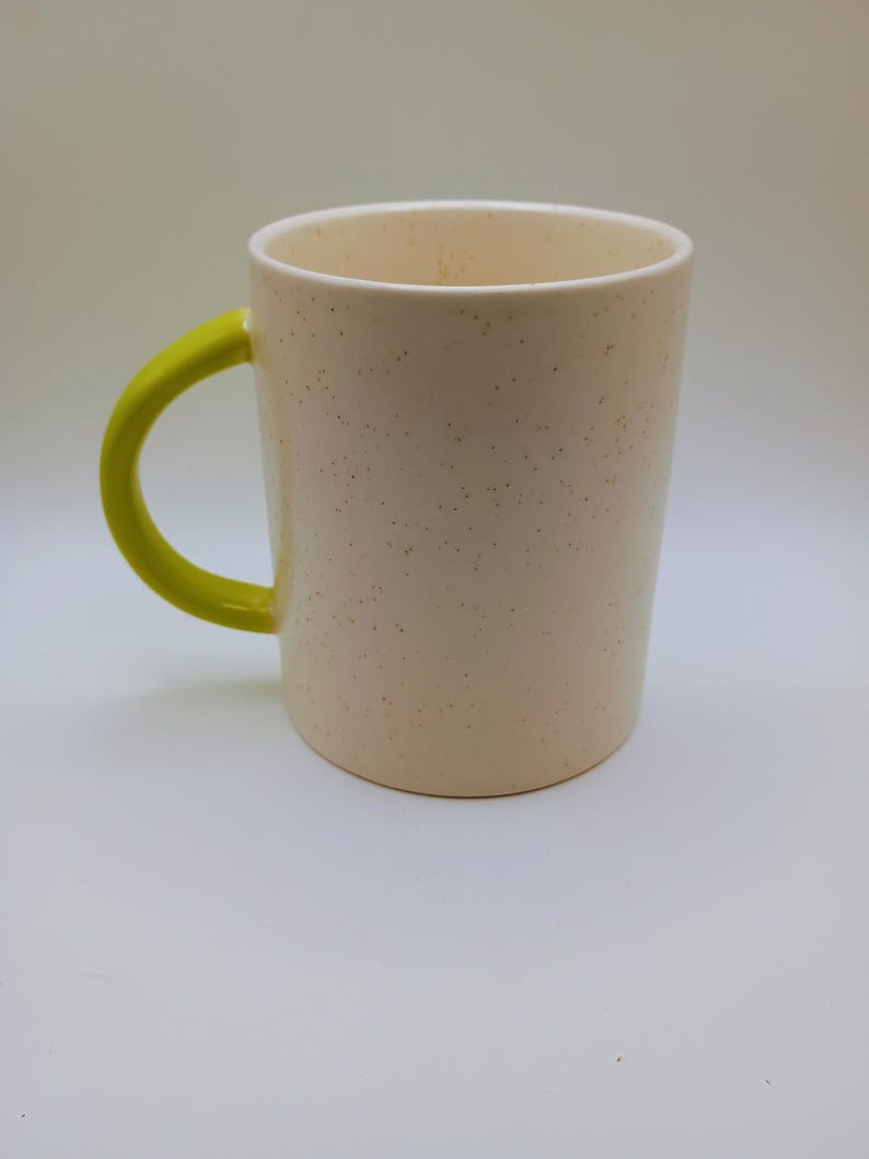 May include: A speckled cream-colored ceramic mug with a bright yellow handle. The mug has a cylindrical shape and a simple design, suitable for coffee, tea, or other beverages. The interior of the mug is white.