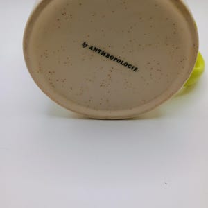 May include: Cream-colored ceramic mug with a speckled base and a bright green handle. The words "by ANTHROPOLOGIE" are printed in black on the bottom of the mug. A bright green stripe is visible on the side of the mug.
