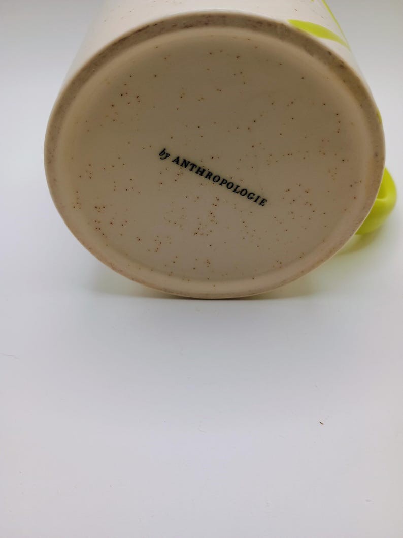 May include: Cream-colored ceramic mug with a speckled base and a bright green handle. The words "by ANTHROPOLOGIE" are printed in black on the bottom of the mug. A bright green stripe is visible on the side of the mug.