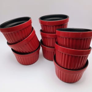 Op de afbeelding: Een verzameling van acht rode ramekins met zwarte binnenkant. De ramekins zijn in groepen gestapeld, wat hun geribbelde buitenkant laat zien. Deze kleine, ovenbestendige schalen zijn ideaal voor het bakken van individuele porties desserts of hartige gerechten.