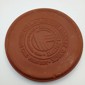 May include: A round, reddish-brown brick pottery lid with embossed text and a central logo. The text reads "BUTTON BASKET BRICK POTTERY" and "CARE WHEN HOT." The logo features a stylized design. Made in Roseville, Ohio.