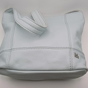 May include: A light blue shoulder bag with a textured surface and visible stitching. The bag features a single strap and a small silver-toned metal logo tag on the lower right side that reads The Sak in a stylized font. The design is structured.