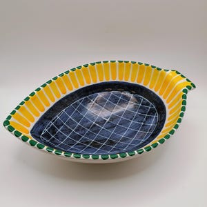 Puede incluir: Una fuente de cerámica con forma de hoja y base blanca. El borde está decorado con detalles amarillos y verdes. El interior presenta un fondo azul oscuro con un patrón de cuadrícula blanca. Una pieza decorativa para la cocina o el comedor.