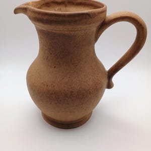 May include: A brown ceramic pitcher with a handle and a spout. The pitcher has a rounded body and a textured surface. The handle is curved and attached to the side of the pitcher. The spout is small and angled. The pitcher is likely used for pouring liquids.