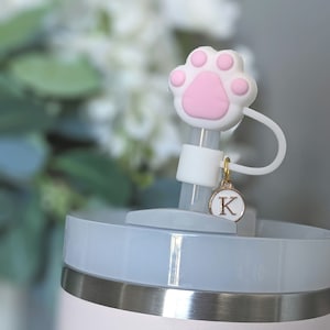 May include: A white and pink paw print straw topper with a gold charm that says "K" on a white tumbler with a light blue lid.