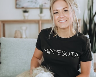 May include: A woman wearing a black t-shirt with the word "MINNESOTA" printed in white on the front. She is sitting on a couch with a small white dog.