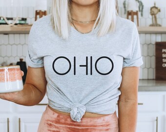 May include: A gray t-shirt with the word "OHIO" printed in black. The shirt is tied at the waist and the model is wearing a pink skirt. The shirt is described as "athletic heather".