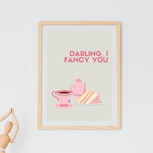 May include: A framed print with a light beige background and pink text that reads "Darling, I fancy you". The print features a pink teacup with a brown liquid inside, a pink teapot, and a slice of pink cake on a white plate.